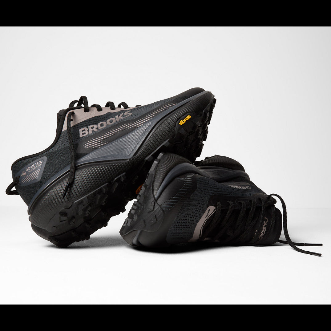 A pair of Brooks mountain biking shoes in black and dark gray, with one shoe standing upright and the other lying on its side to show the sole, on a light gray studio background.
