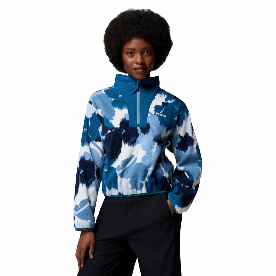 A woman with an afro hairstyle wearing a blue and white abstract patterned quarter-zip pullover and black pants, standing against a solid white background.