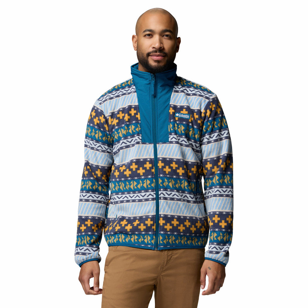 A man wearing a patterned fleece jacket with blue, navy, white, and yellow geometric designs, paired with tan pants, against a plain white background.