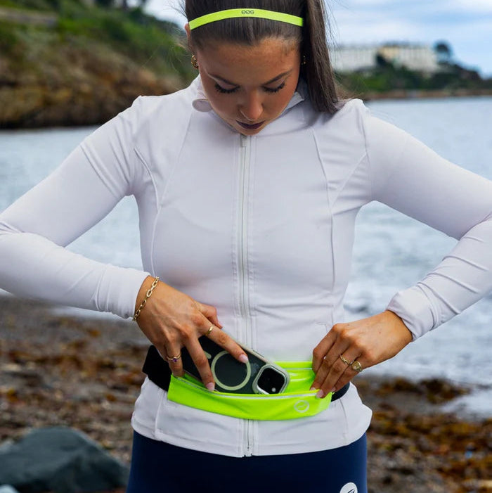 A woman on a beach wearing a white long-sleeve athletic top and dark blue leggings, adjusting a bright neon yellow waist bag that contains a smartphone. She is also wearing a matching neon yellow headband.