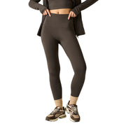 A model wearing high-waisted dark brown athletic leggings, styled with a matching dark brown zip-up jacket, beige socks, and brown and beige sneakers, set against a plain white background.