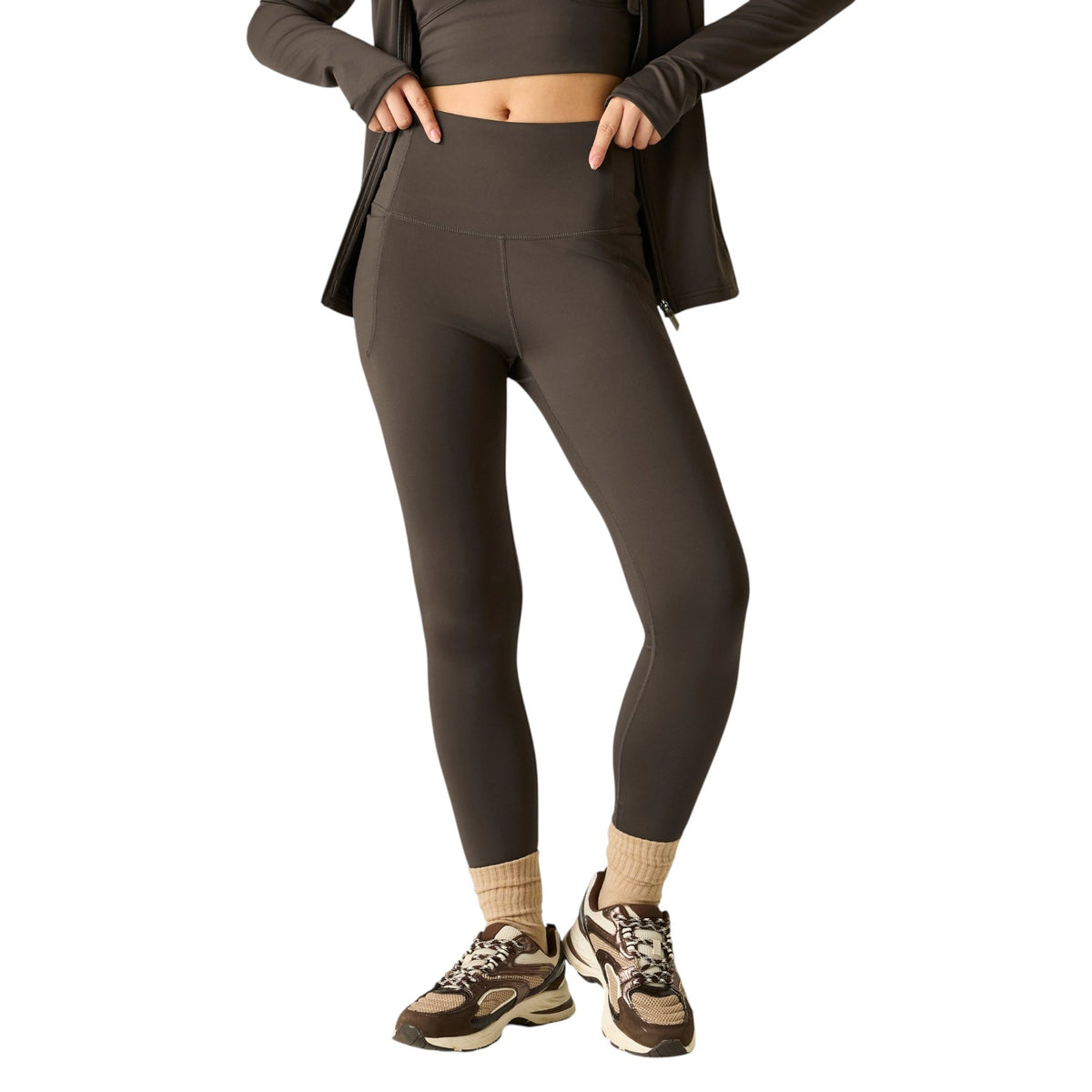 A model wearing high-waisted dark brown athletic leggings, styled with a matching dark brown zip-up jacket, beige socks, and brown and beige sneakers, set against a plain white background.