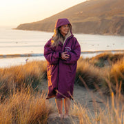 A woman wearing a purple hooded beach poncho stands on a sandy path in a coastal grassy area at sunset, with the ocean and hills in the background.