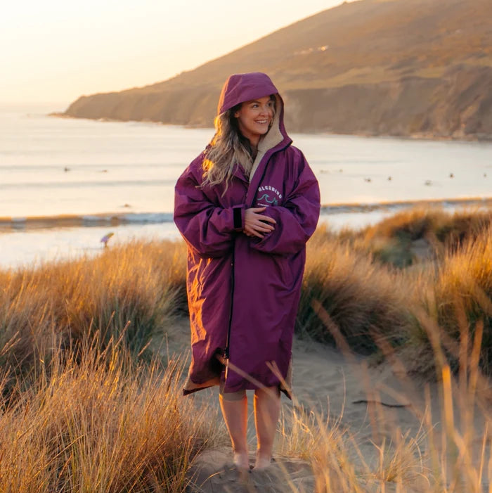 A woman wearing a purple hooded beach poncho stands on a sandy path in a coastal grassy area at sunset, with the ocean and hills in the background.